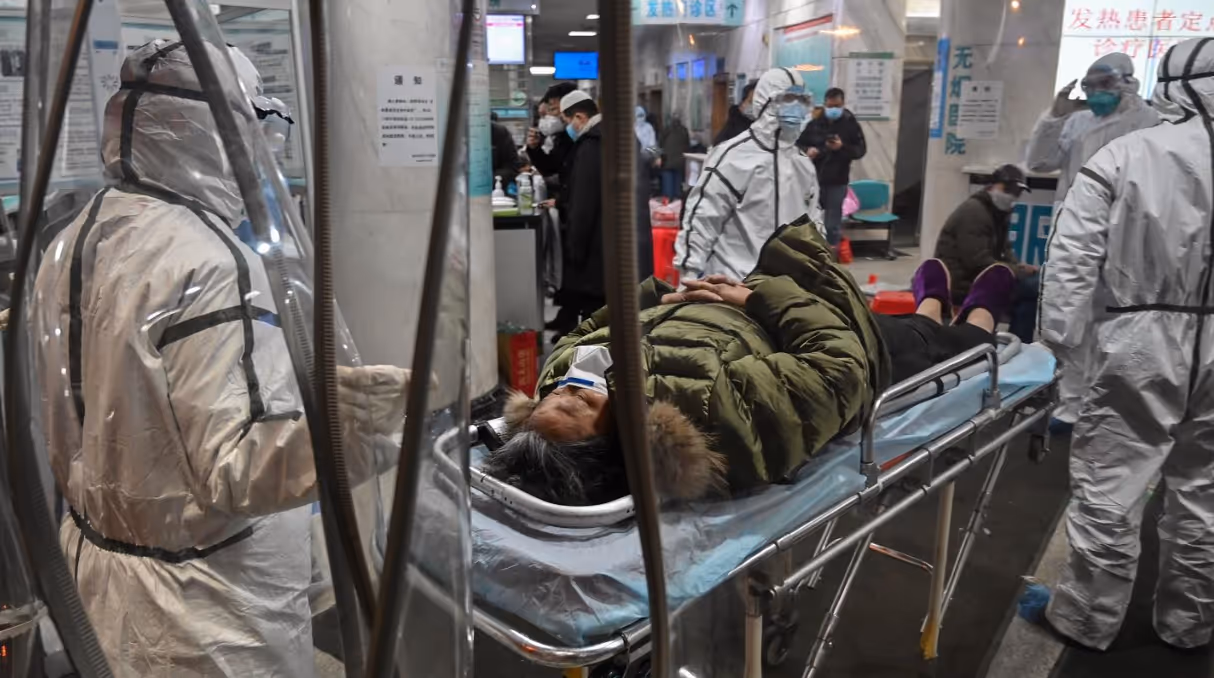 Person wearing a green jacket and face mask lying on a hospital gurney surrounded by medical staff in full protective suits.